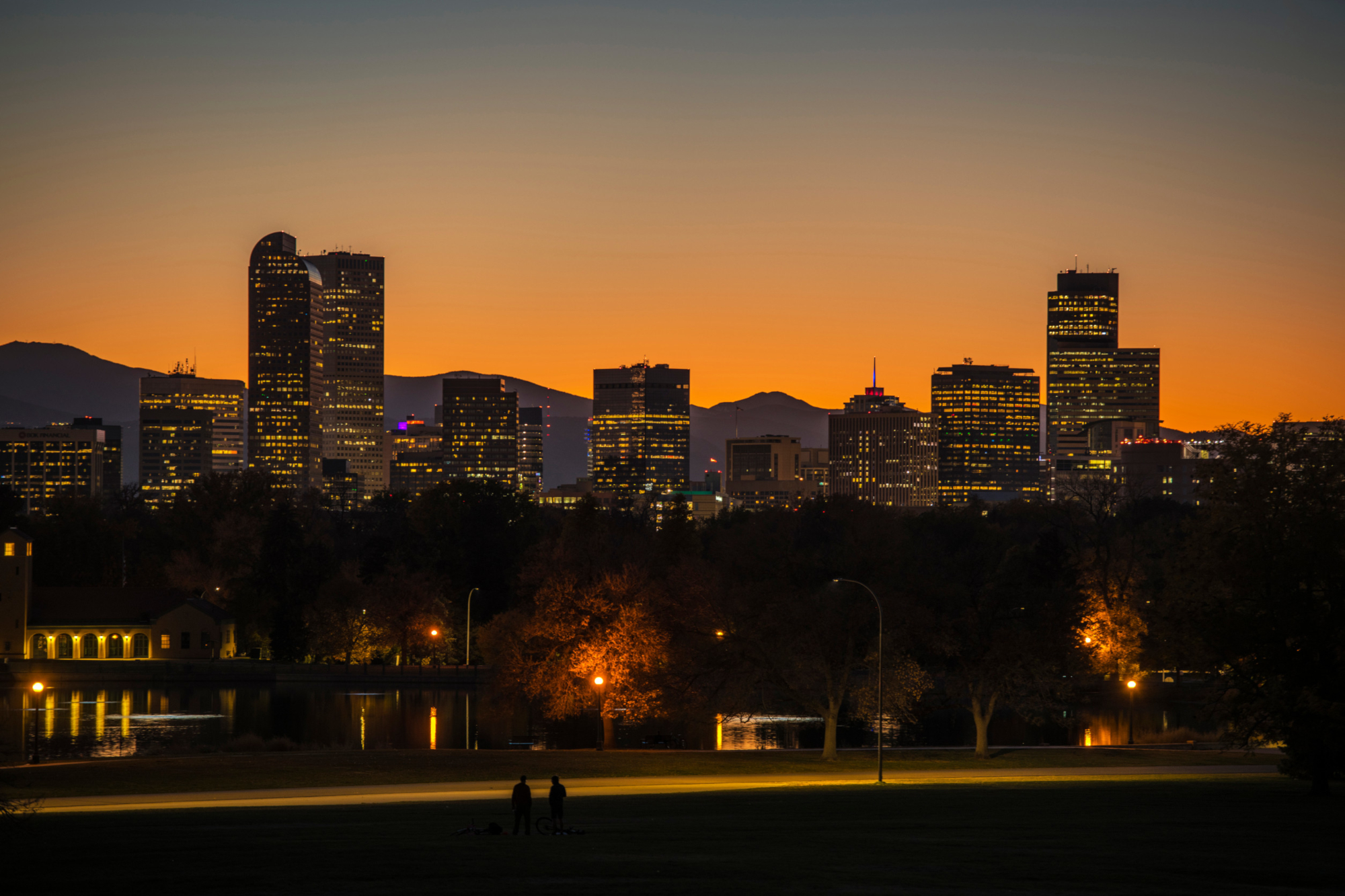 Denver city skyline at sunset with buildings silhouetted against an orange sky, trees and streetlights visible in the foreground, and mountains in the background.