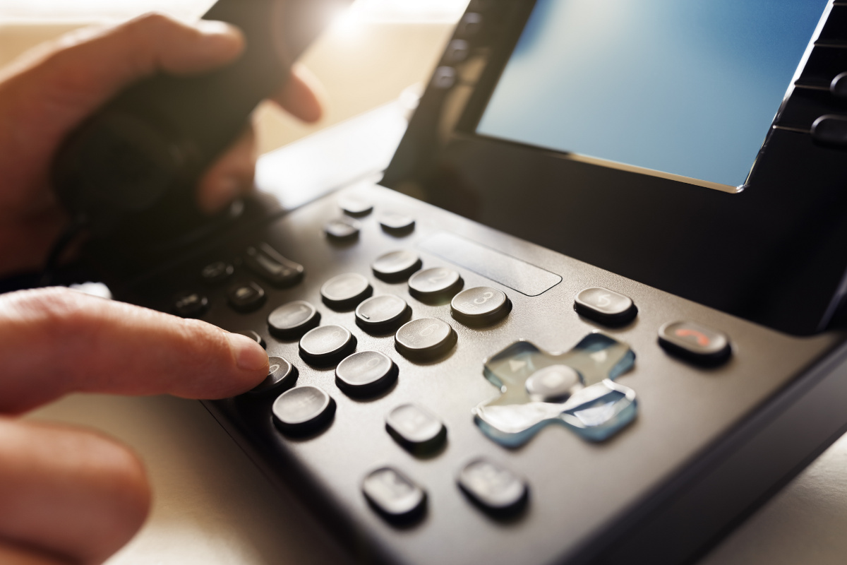 Close up of an office phone in use, with one hand dialing on the keypad and the other holding the receiver out of focus in the background.