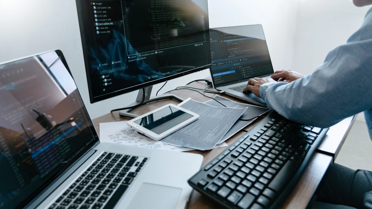 A man seated at a desk, focused on three computer monitors displaying various tasks and information.